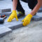 Buttrose Landscaping - person in yellow gloves lays paving stones in layers. Garden brick pathway paving by professional paver worker. Laying gray concrete paving slabs in house courtyard on sand foundation base.